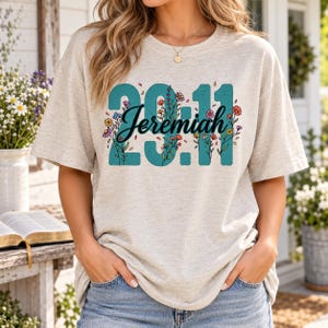 May include: A heathered beige short-sleeve t-shirt featuring the teal text 29:11 with the word Jeremiah written in black cursive script across the center. The design is decorated with colorful floral illustrations intertwined with the numbers.