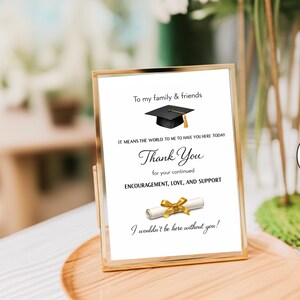 May include: A vertical rectangular gold-toned metal frame containing a white sign with black text that reads: To my family & friends, IT MEANS THE WORLD TO ME TO HAVE YOU HERE TODAY, Thank You, for your continued ENCOURAGEMENT, LOVE, AND SUPPORT, I wouldn't be here without you! The sign features a black graduation cap with a gold tassel and a white diploma scroll tied with a gold ribbon.