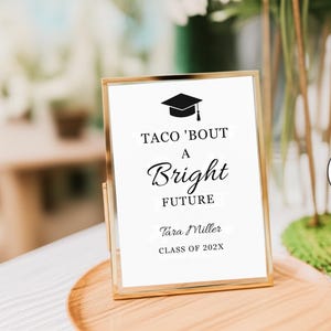 May include: A vertical rectangular sign in a thin gold metal frame featuring a black graduation cap icon and the text TACO 'BOUT A BRIGHT FUTURE, Tara Miller, and CLASS OF 202X printed in black ink on a white background.