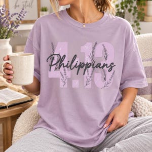 May include: A light purple short-sleeve t-shirt featuring the text 4.13 and the word Philippians in black cursive script. The numbers are filled with a light lavender pattern of sprigs and stems. The shirt has a crew neckline and a relaxed fit.