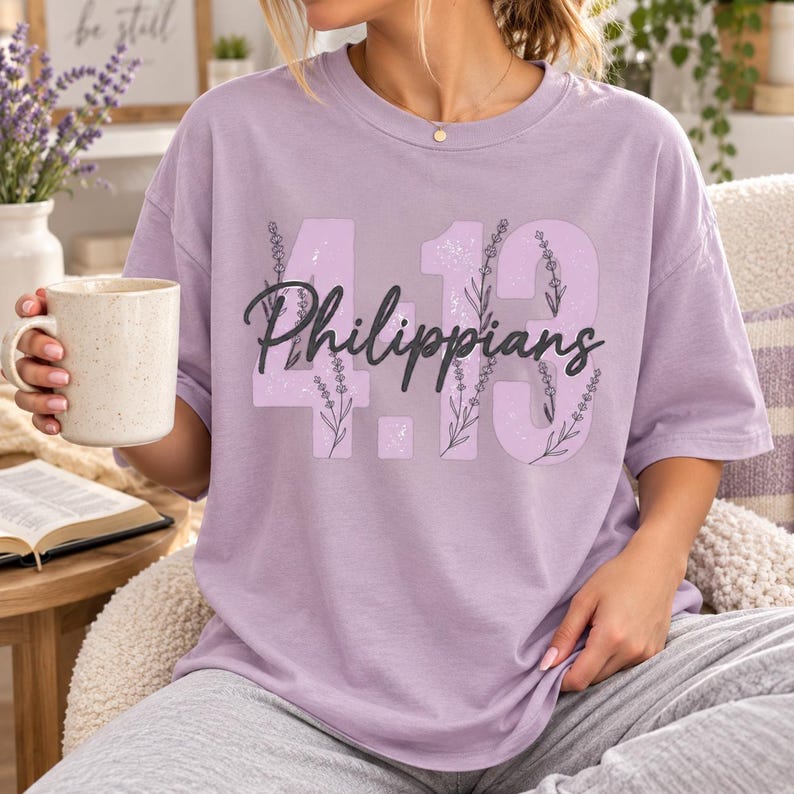 May include: A light purple short-sleeve t-shirt featuring the text 4.13 and the word Philippians in black cursive script. The numbers are filled with a light lavender pattern of sprigs and stems. The shirt has a crew neckline and a relaxed fit.