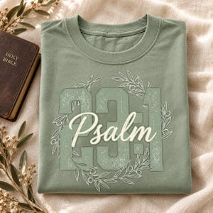 May include: A sage green t-shirt featuring a distressed graphic design with the large numbers 23:1, the word Psalm in a white script font, and an olive branch wreath encircling the text. The shirt has a crew neckline and short sleeves.