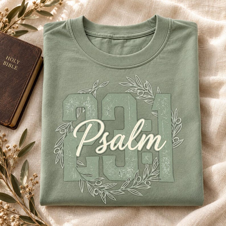 May include: A sage green t-shirt featuring a distressed graphic design with the large numbers 23:1, the word Psalm in a white script font, and an olive branch wreath encircling the text. The shirt has a crew neckline and short sleeves.
