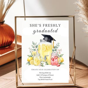 May include: A rectangular sign in a gold frame featuring the text SHE'S FRESHLY graduated and CREATE YOUR CELEBRATION SIP. The illustration shows a large lemonade dispenser with a graduation cap, two mason jars, lemons, and strawberries.