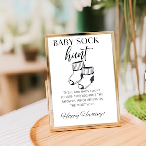 May include: A framed sign with the text "BABY SOCK hunt" in black script. Below the text is an illustration of two baby socks. The sign reads "THERE ARE BABY SOCKS HIDDEN THROUGHOUT THE SHOWER. WHOEVER FINDS THE MOST WINS! Happy Hunting!"