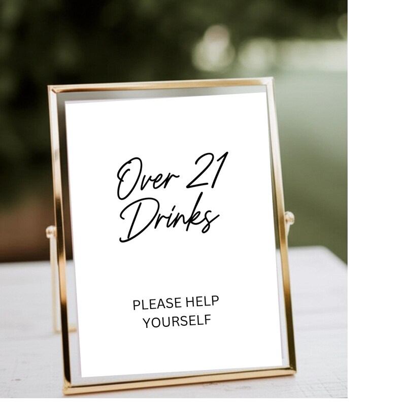 Op de afbeelding: Een goudkleurig omlijst bord met de tekst "Over 21 Drinks" in elegant schrift. Daaronder staat "PLEASE HELP YOURSELF". Het bord staat tegen een vage groene achtergrond, wat duidt op een buitenevenement of bijeenkomst.