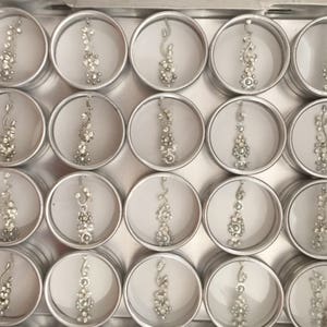 Op de afbeelding: Een verzameling van 20 kleine zilveren containers met decoratieve zilveren bindi-stickers erin. De containers zijn gerangschikt in een rasterpatroon op een zilveren dienblad.