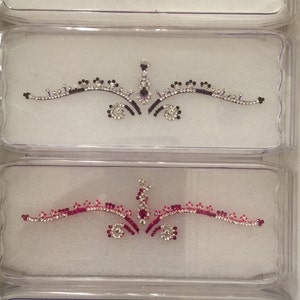 May include: Four clear plastic cases each containing a decorative adhesive forehead bindi. Each bindi features a symmetrical design of clear rhinestones accented with red, purple, magenta, or dark red colored stones in floral and swirl patterns.