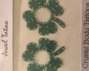 Puede incluir: Dos tatuajes de joyas en forma de trébol verde con diamantes de imitación transparentes. Los tatuajes están sobre un fondo blanco con el texto "Jewel Tattoo" y "Crystal Body Tattoos" en los lados izquierdo y derecho de la imagen.