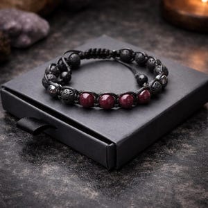 Könnte beinhalten: Ein schwarzes Perlenarmband mit schwarzen Lavasteinperlen und tiefroten Edelsteinperlen. Das Armband wird auf einer schwarzen Schachtel präsentiert, die den Kontrast hervorhebt. Das Armband ist verstellbar.