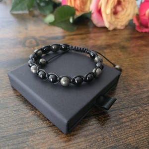 May include: A black beaded bracelet with pyrite and onyx stones, presented on a black box. The bracelet has a woven design and an adjustable cord. The background features roses on a wooden surface.