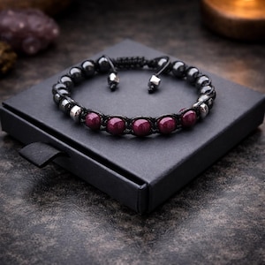 May include: A black beaded bracelet with dark red and silver accents, resting on a black box. The bracelet features dark gray beads and a series of dark red, round beads. The bracelet is adjustable with a sliding knot closure.
