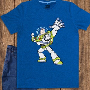 May include: A blue t-shirt featuring a cartoon image of Buzz Lightyear from Toy Story. The character is depicted in his signature white, green, and purple spacesuit, with his arm raised. A pair of blue jeans is next to the shirt.