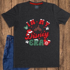 Może przedstawiać: Ciemnoszara koszulka z napisem "IN MY Disney ERA" w kolorze czerwonym i zielonym, z grafiką Myszki Miki i płatk&oacute;w śniegu. Koszulka jest zestawiona z niebieskimi dżinsami na drewnianej powierzchni.