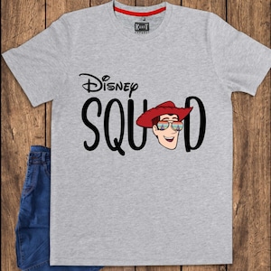 May include: A heather gray short-sleeve t-shirt featuring the black text Disney SQUAD, with the letter U replaced by a cartoon illustration of a character wearing a red cowboy hat and sunglasses reflecting a castle and sunset landscape.