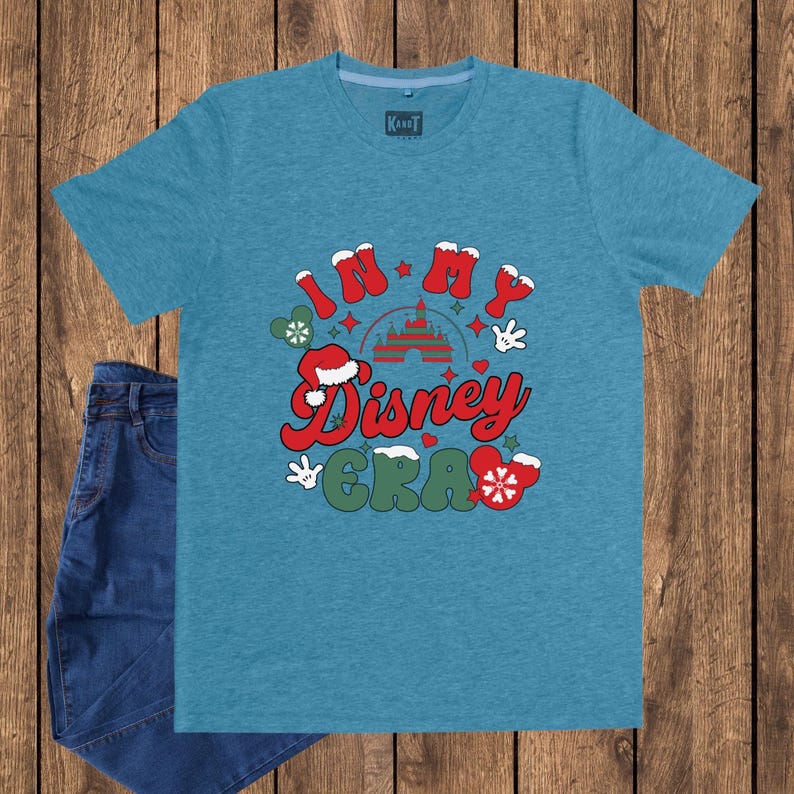 Może przedstawiać: Turkusowa koszulka z napisem "IN MY Disney ERA" w kolorze czerwonym, zielonym i białym. Projekt zawiera rękawice Myszki Miki, czapkę Świętego Mikołaja i zamek. Obok koszulki znajduje się para niebieskich dżins&oacute;w.