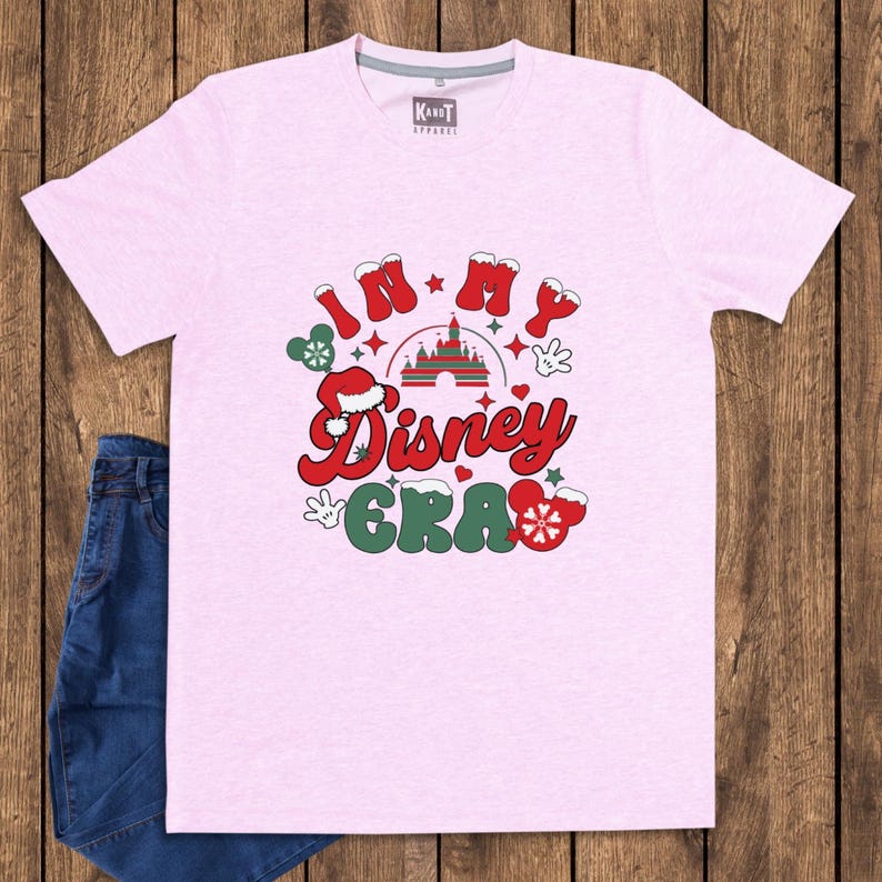 Może przedstawiać: Jasnor&oacute;żowa koszulka z napisem "IN MY Disney ERA" w kolorze czerwonym i zielonym. Projekt zawiera zamek, płatki śniegu i dłonie Myszki Miki. Koszulka jest zestawiona z niebieskimi dżinsami na drewnianym tle.
