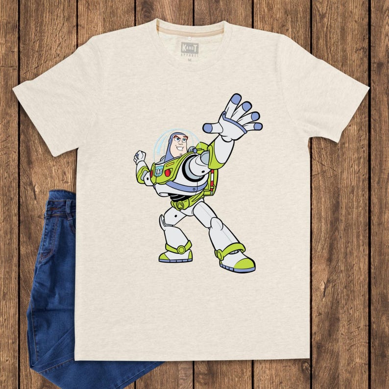 May include: A light beige t-shirt featuring a cartoon illustration of Buzz Lightyear from Toy Story. The character is depicted in his signature spacesuit, with green, white, and blue accents. Blue jeans are visible on the left side of the image.