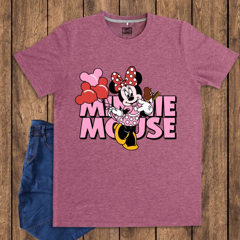 May include: A maroon t-shirt featuring a graphic of Minnie Mouse holding balloons and a treat, with the words "Minnie Mouse" in pink. The shirt is paired with blue jeans, displayed on a wooden surface.