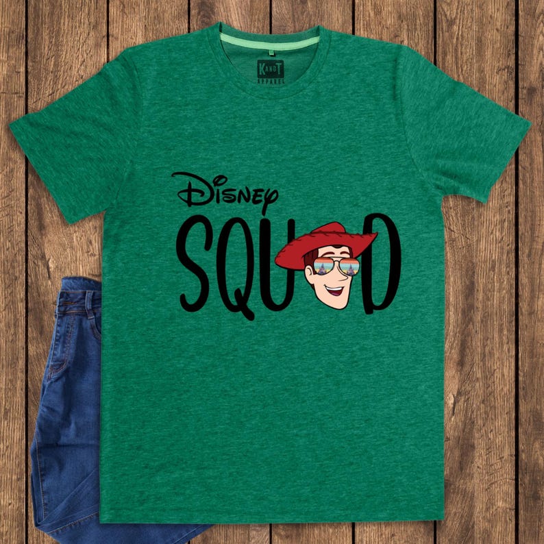 May include: A heather green short-sleeve t-shirt featuring the black text Disney SQUAD with a graphic of a cartoon character wearing a red cowboy hat and sunglasses. The shirt has a crew neckline and a small interior label reading KANDT APPAREL.