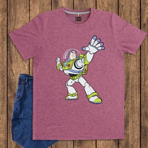 May include: A maroon t-shirt featuring a cartoon illustration of Buzz Lightyear in his spacesuit. The graphic shows Buzz with his arm raised. A pair of blue jeans is folded next to the shirt.