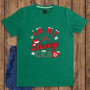 Może przedstawiać: Zielona koszulka z napisem "IN MY Disney ERA" w kolorze czerwonym i białym, z grafiką w stylu Myszki Miki. Koszulka jest połączona z niebieskimi dżinsami, na tle drewnianego tła.