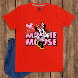 May include: A red t-shirt featuring a graphic of Minnie Mouse holding balloons and a treat. The text "MINNIE MOUSE" is displayed in pink and black. A pair of blue jeans is visible on the left side of the image.