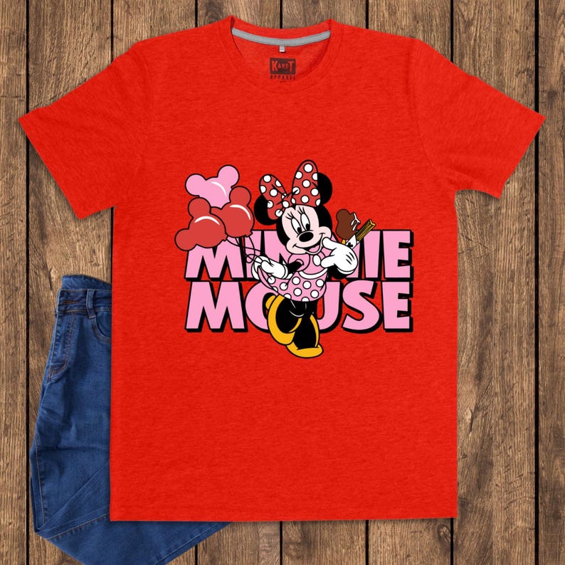 May include: A red t-shirt featuring a graphic of Minnie Mouse holding balloons and a treat. The text "MINNIE MOUSE" is displayed in pink and black. A pair of blue jeans is visible on the left side of the image.