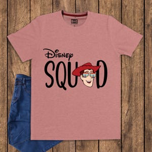 May include: A dusty rose short-sleeved t-shirt featuring the black text Disney SQUAD with a graphic of a cowboy character wearing a red hat and sunglasses reflecting a castle scene. The shirt has a crew neckline and a small interior brand label.