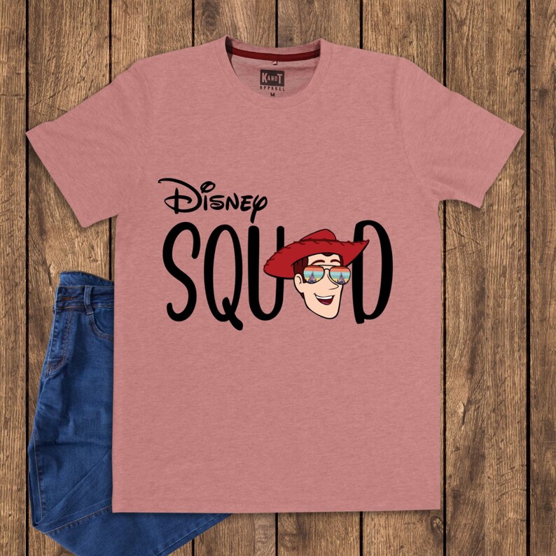 May include: A dusty rose short-sleeved t-shirt featuring the black text Disney SQUAD with a graphic of a cowboy character wearing a red hat and sunglasses reflecting a castle scene. The shirt has a crew neckline and a small interior brand label.