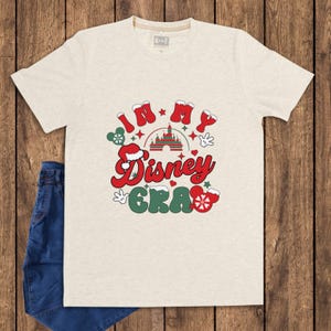 Może przedstawiać: Jasny beżowy t-shirt z napisem "IN MY Disney ERA" w kolorze czerwonym i zielonym, z uszami Myszki Miki i grafiką zamku. Koszulka jest połączona z niebieskimi dżinsami na drewnianym tle.