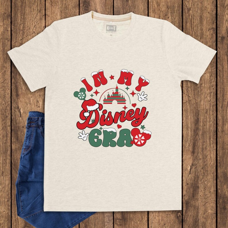 Może przedstawiać: Jasny beżowy t-shirt z napisem "IN MY Disney ERA" w kolorze czerwonym i zielonym, z uszami Myszki Miki i grafiką zamku. Koszulka jest połączona z niebieskimi dżinsami na drewnianym tle.