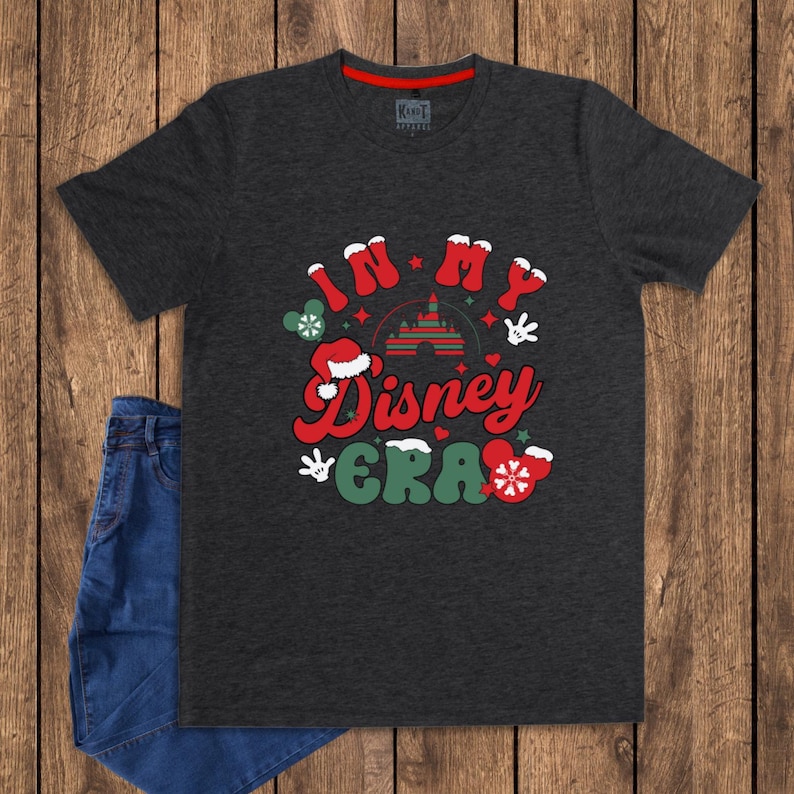 Może przedstawiać: Ciemnoszara koszulka z napisem "IN MY Disney ERA" w kolorze czerwonym i zielonym, z grafiką Myszki Miki i płatk&oacute;w śniegu. Koszulka jest zestawiona z niebieskimi dżinsami na drewnianej powierzchni.