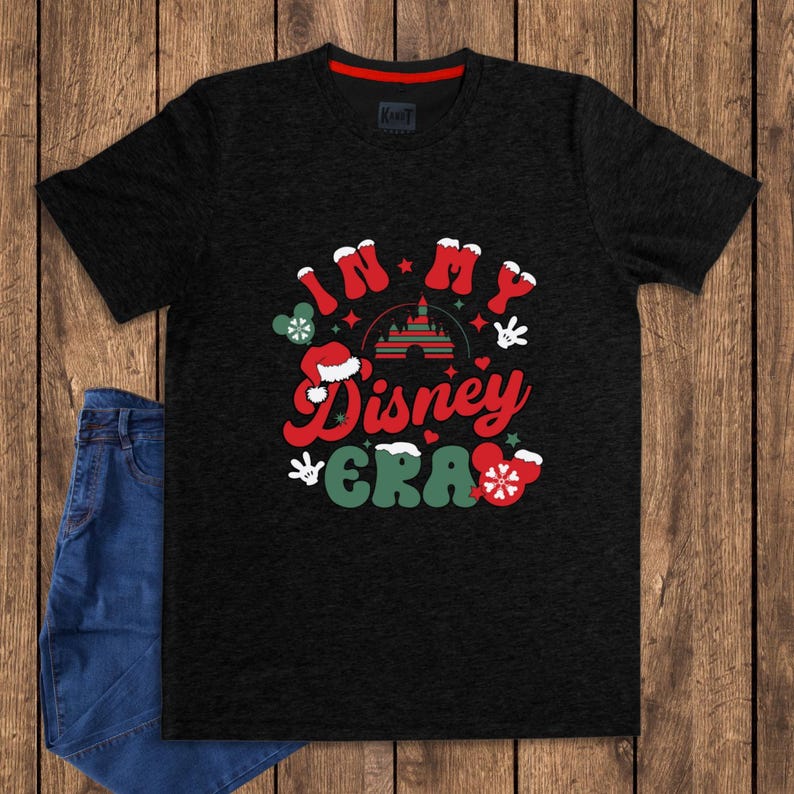 Może przedstawiać: Czarny t-shirt z napisem "In My Disney Era" w kolorze czerwonym i zielonym, ozdobiony świątecznymi grafikami Disneya. Projekt zawiera zamek, rękawice Myszki Miki i czapkę Świętego Mikołaja. Koszulka ma czerwony kołnierz.