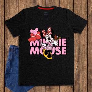 May include: Black t-shirt featuring Minnie Mouse holding balloons and a chocolate treat. The text "MINNIE MOUSE" is in pink. The shirt has a red collar and is paired with blue jeans.