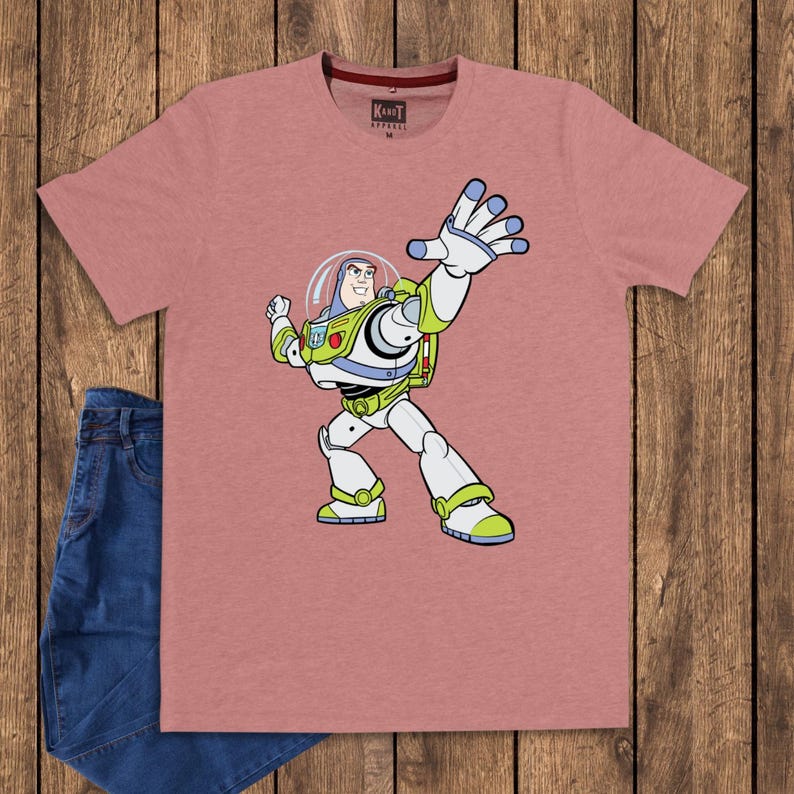 May include: A dusty rose t-shirt featuring a cartoon image of Buzz Lightyear from Toy Story. The character is depicted in his signature white, green, and purple spacesuit, with his arm outstretched. The shirt is paired with blue jeans.