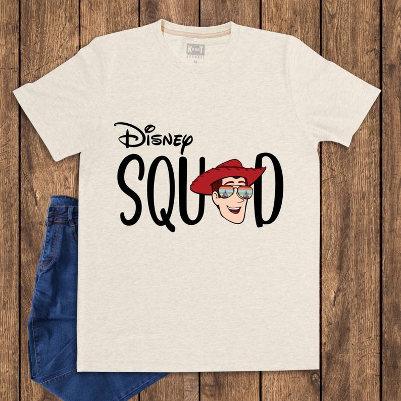 May include: A heathered beige short-sleeve t-shirt featuring the black text Disney SQUAD. The letter U is replaced by a graphic of a cartoon character wearing a red cowboy hat and sunglasses with a colorful sunset reflection on the lenses.