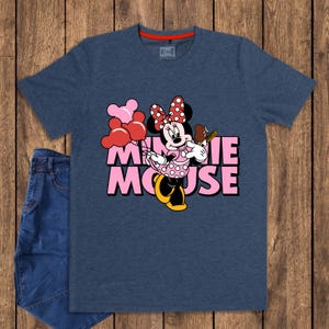 May include: A navy blue t-shirt featuring Minnie Mouse holding balloons and a treat. The graphic includes the text "MINNIE MOUSE" in pink. The shirt is paired with blue jeans on a wooden background.