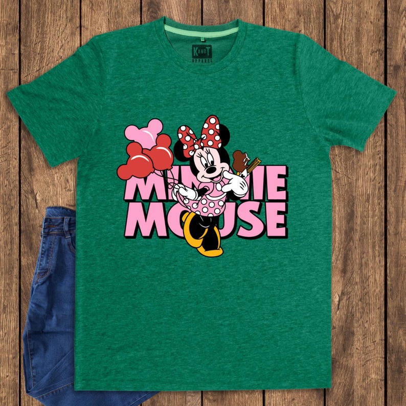 May include: Green t-shirt featuring a graphic of Minnie Mouse holding balloons and a chocolate bar. The text "MINNIE MOUSE" is in pink. The shirt is paired with blue denim jeans. The brand "KARAT APPAREL" is printed on the shirt.
