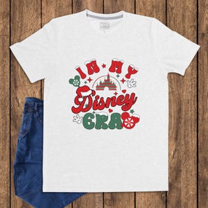 Może przedstawiać: Jasnoszara koszulka z napisem "In My Disney Era" w kolorze czerwonym i zielonym, z grafiką zamku i ikonami Myszki Miki. Koszulka jest połączona z niebieskimi dżinsami na drewnianym tle.