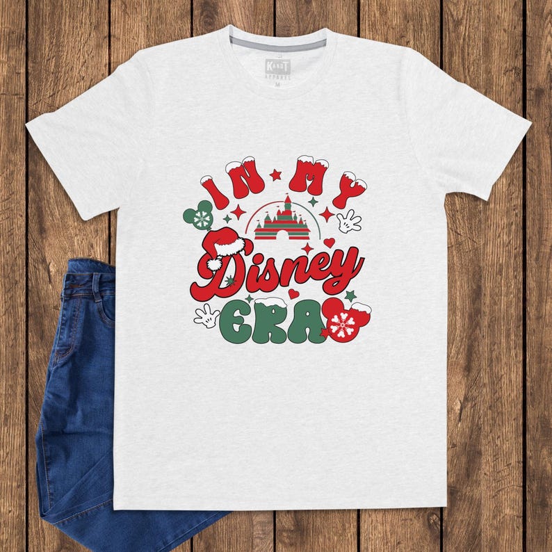 Może przedstawiać: Jasnoszara koszulka z napisem "In My Disney Era" w kolorze czerwonym i zielonym, z grafiką zamku i ikonami Myszki Miki. Koszulka jest połączona z niebieskimi dżinsami na drewnianym tle.