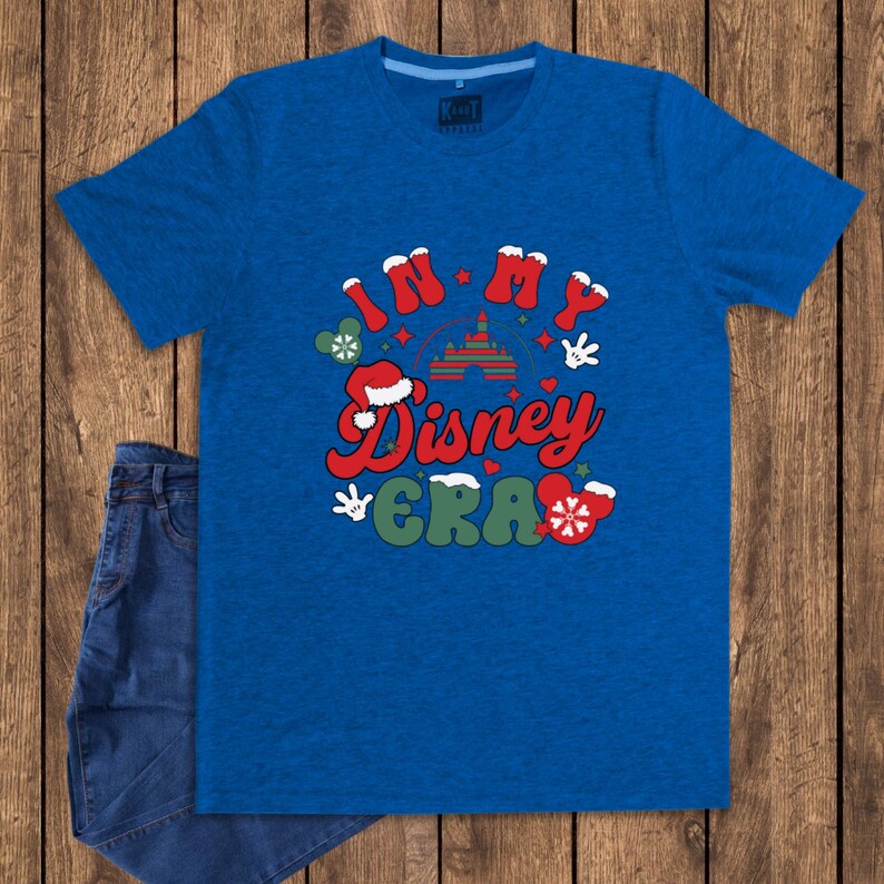 Może przedstawiać: Niebieska koszulka z napisem "IN MY Disney ERA" w kolorze czerwonym, zielonym i białym. Projekt zawiera rękawice Myszki Miki, czapkę Świętego Mikołaja i sylwetkę zamku. Para niebieskich dżins&oacute;w jest widoczna po lewej stronie obrazu.