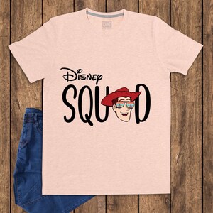 May include: A light peach-colored short-sleeve t-shirt featuring the black text Disney SQUAD. The letter U is replaced by a graphic of a cartoon character wearing a red cowboy hat and sunglasses with a colorful sunset reflection on the lenses.