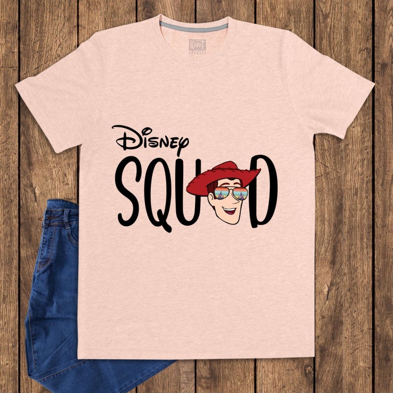 May include: A light peach-colored short-sleeve t-shirt featuring the black text Disney SQUAD. The letter U is replaced by a graphic of a cartoon character wearing a red cowboy hat and sunglasses with a colorful sunset reflection on the lenses.