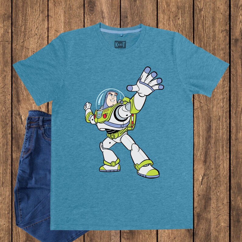 May include: A teal t-shirt featuring a cartoon illustration of Buzz Lightyear from Toy Story. The character is depicted in his signature spacesuit, with white, green, and purple accents. A pair of blue jeans is visible on the left side of the image.