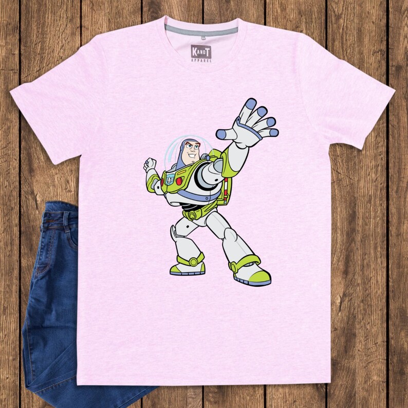 May include: A light pink t-shirt featuring a graphic of Buzz Lightyear from Toy Story. The cartoon character is in a dynamic pose with his arm raised. The shirt is paired with blue jeans, suggesting casual wear. The shirt has the text "KandT Apparel" on the tag.