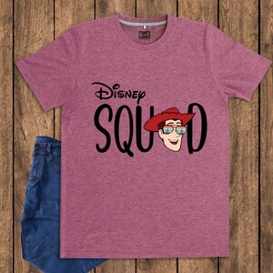 May include: A heathered mauve short-sleeve t-shirt featuring the black text Disney SQUAD. The letter U is replaced by a graphic of a smiling character wearing a red cowboy hat and sunglasses with a colorful sunset reflection on the lenses.