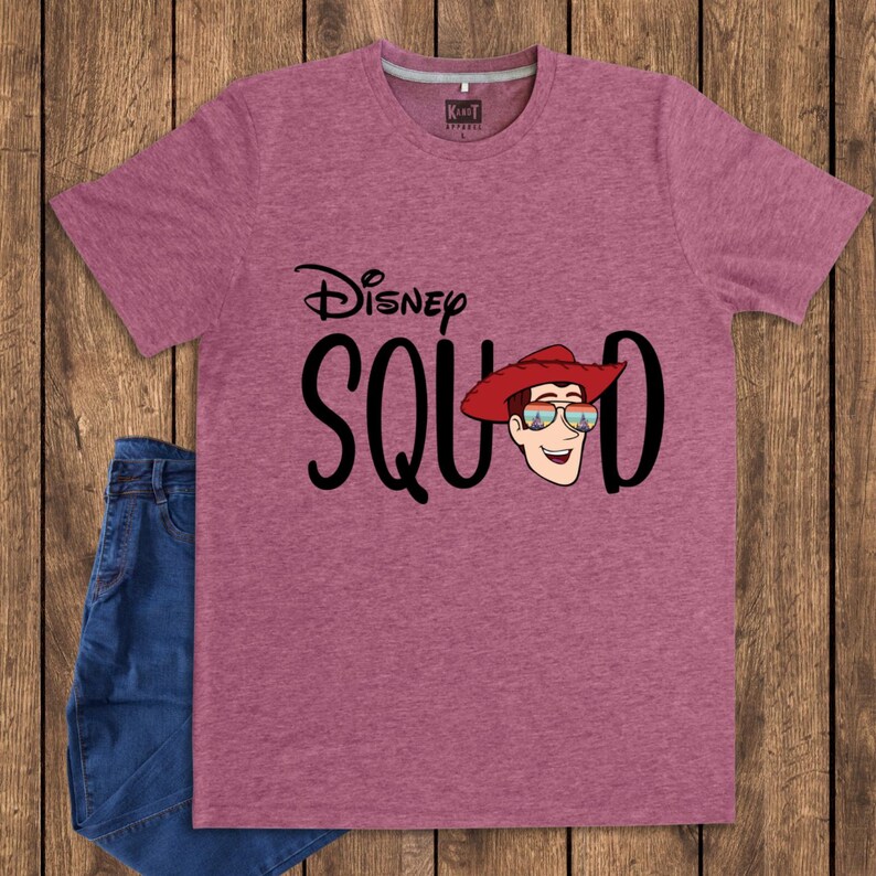May include: A heathered mauve short-sleeve t-shirt featuring the black text Disney SQUAD. The letter U is replaced by a graphic of a smiling character wearing a red cowboy hat and sunglasses with a colorful sunset reflection on the lenses.