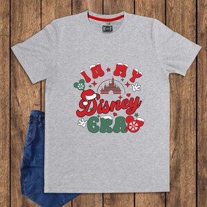 Może przedstawiać: Szara koszulka z napisem "IN MY Disney ERA" w kolorze czerwonym i zielonym, z uszami Myszki Miki, czapką Świętego Mikołaja i grafiką zamku. Koszulka jest połączona z niebieskimi dżinsami.