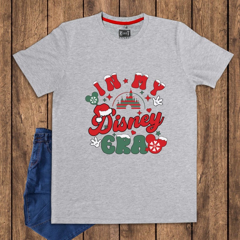 Może przedstawiać: Szara koszulka z napisem "IN MY Disney ERA" w kolorze czerwonym i zielonym, z uszami Myszki Miki, czapką Świętego Mikołaja i grafiką zamku. Koszulka jest połączona z niebieskimi dżinsami.